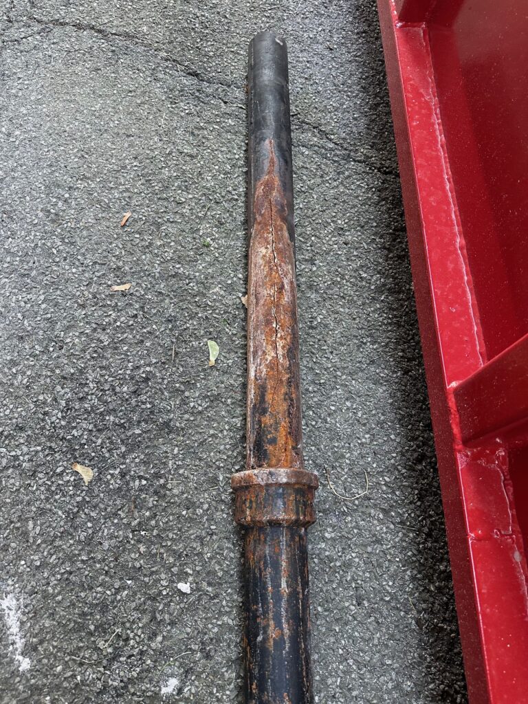Section of old cast iron drain pipe showing heavy rust and a visible crack along its length, resting on pavement