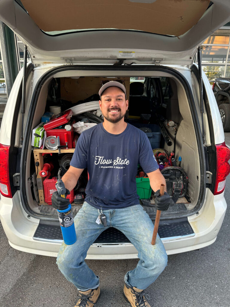 Plumber sitting on the back of a work van holding a torch and copper pipe, wearing a Flow State Plumbing & Drains shirt with tools organized behind him.