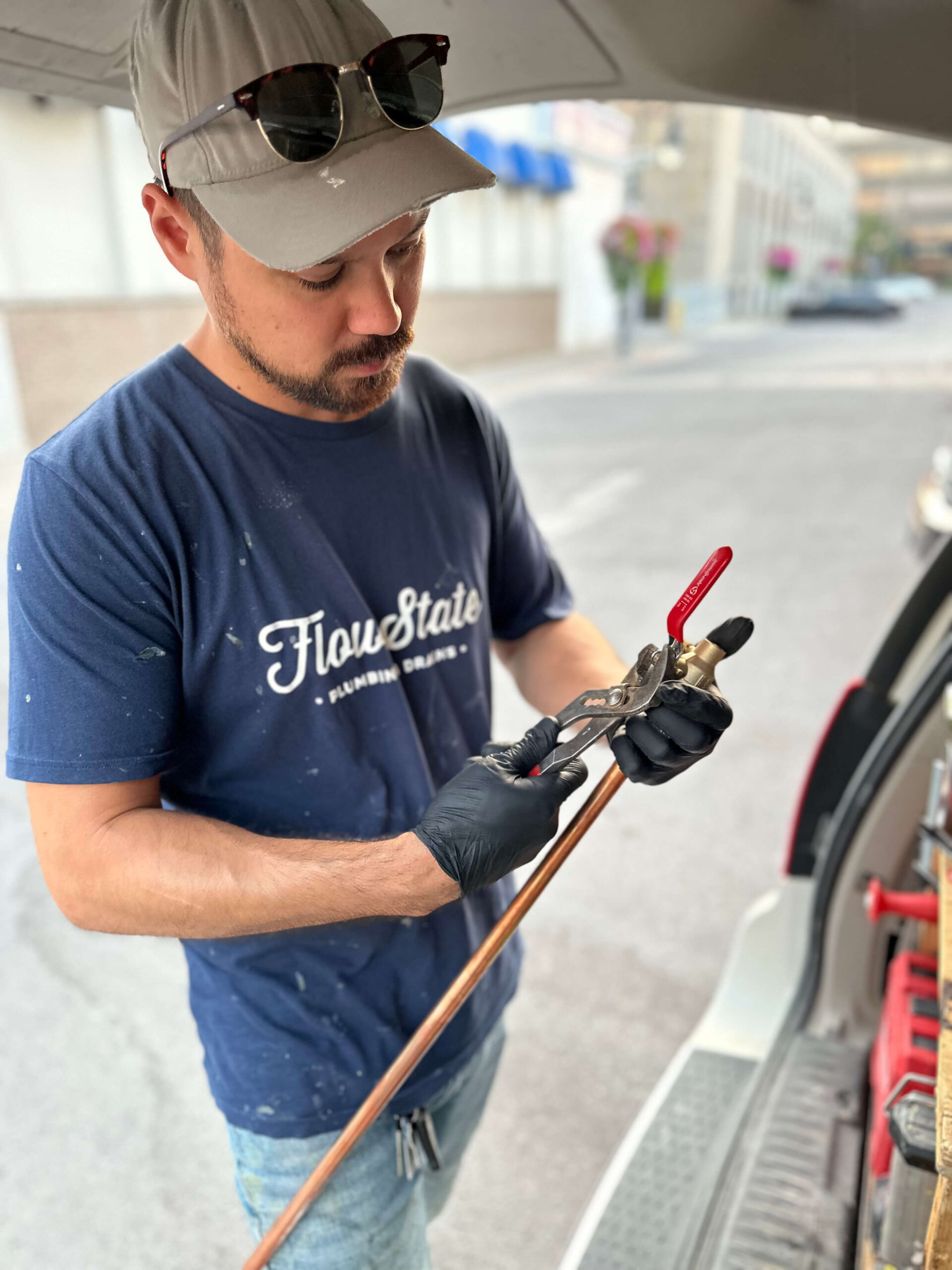Plumber attaching a shut-off valve to a copper pipe using pliers, wearing gloves and Flow State Plumbing & Drains gear.