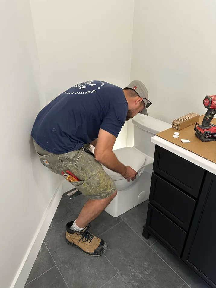 Flow-State Plumbing and Drains technician installing a new modern toilet in a Niagara Region bathroom, with tools and a Milwaukee drill on the vanity beside him.